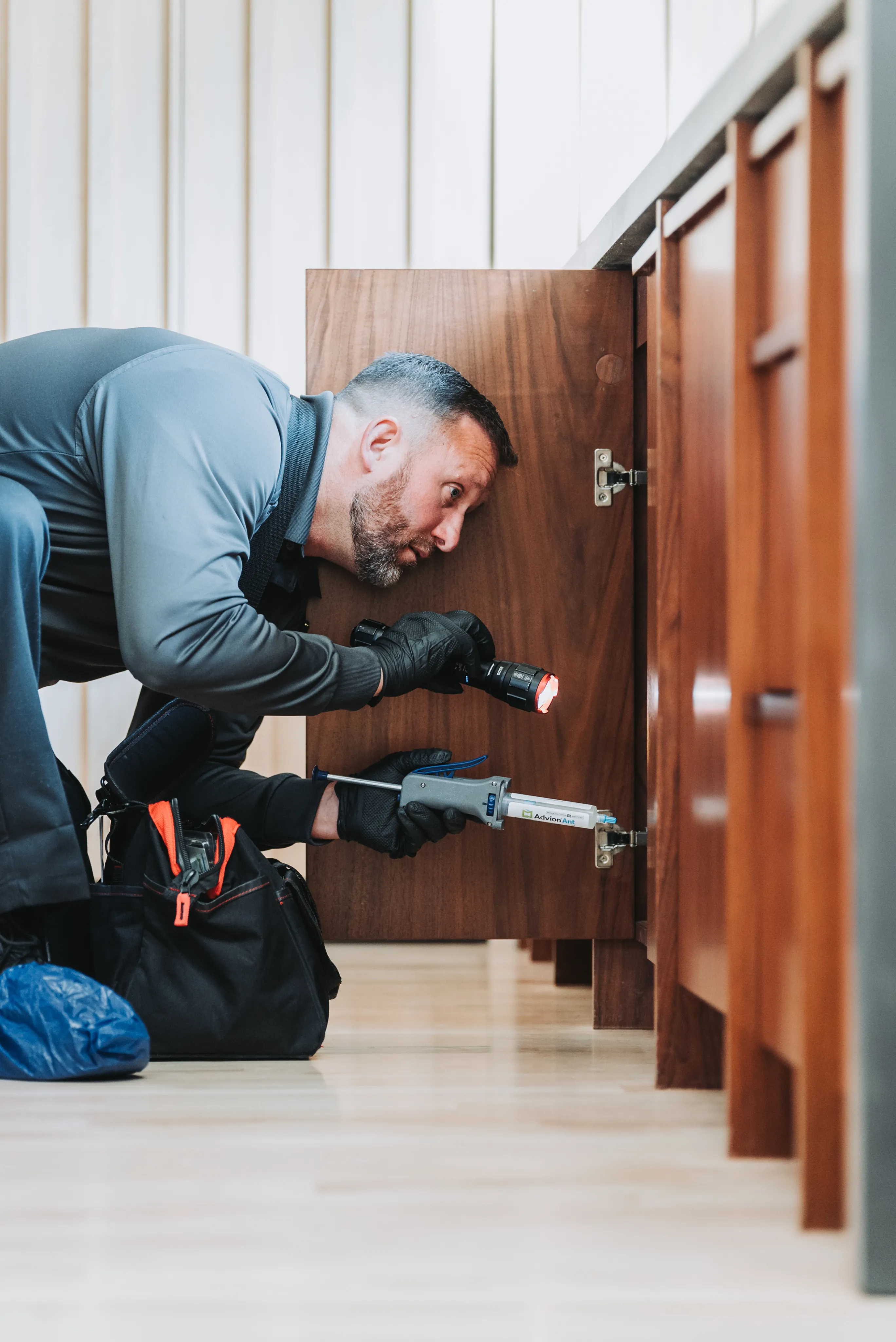 An A&A Pest Control technician kneeling at an open kitchen cabinet with a flashlight and a bait applicator